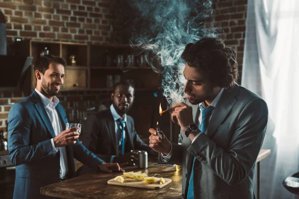 Jeune homme en costume fumant cigare et amis multiethniques buvant des boissons alcoolisées derrière — Photo de stock