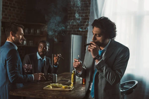 Bonito jovem empresário fumar charuto enquanto amigos beber uísque atrás — Fotografia de Stock