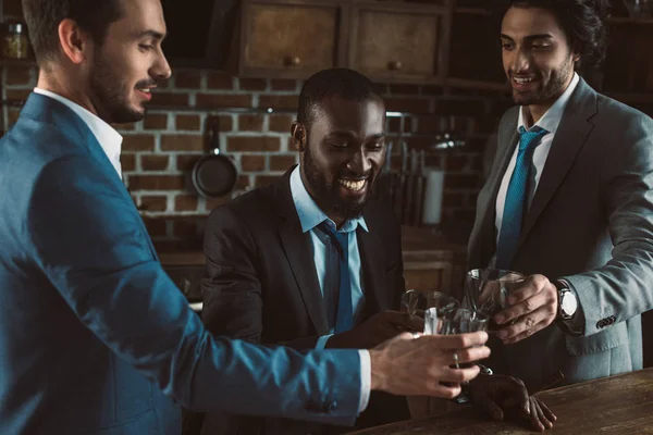 Lächelnde junge multiethnische männliche Freunde in Anzügen klimpern mit Gläsern Whiskey — Stockfoto