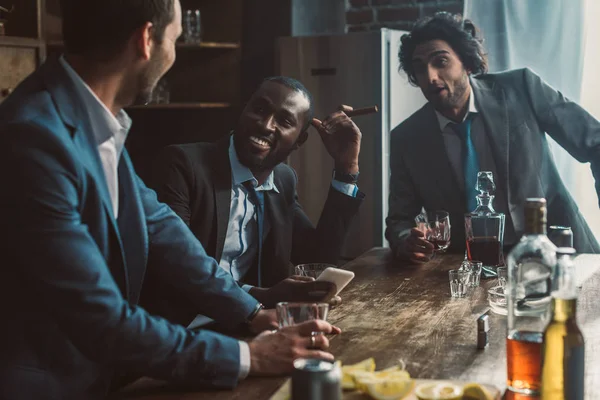 Sonrientes hombres multiétnicos hablando, bebiendo whisky y fumando cigarros mientras festejan juntos - foto de stock