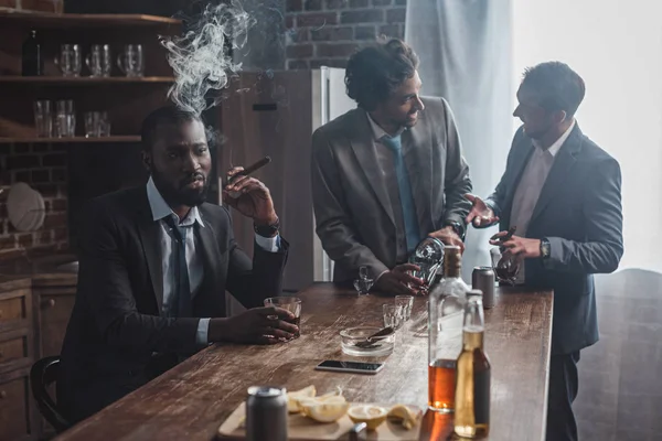 Tres hombres multiétnicos en trajes bebiendo bebidas alcohólicas y fumando cigarros juntos - foto de stock