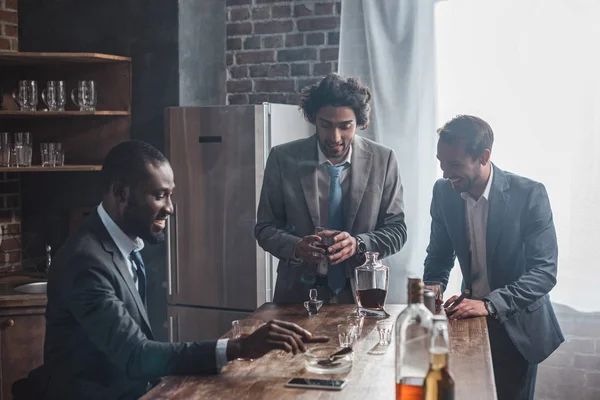 Lächelnde junge multiethnische männliche Freunde in offizieller Kleidung trinken Whiskey und rauchen gemeinsam Zigarren — Stockfoto