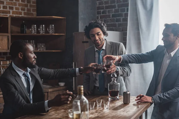 Улыбающиеся многонациональные друзья мужчины звонят в стаканы виски и тусуются вместе — стоковое фото