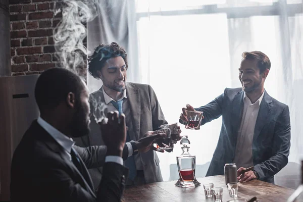 Tres hombres de negocios multiétnicos sonrientes fumando cigarros y tintineando vasos de whisky - foto de stock