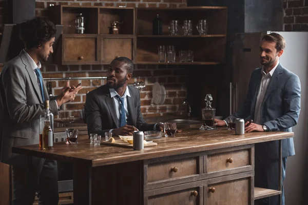 Tres amigos masculinos multiétnicos en trajes hablando mientras beben alcohol juntos - foto de stock