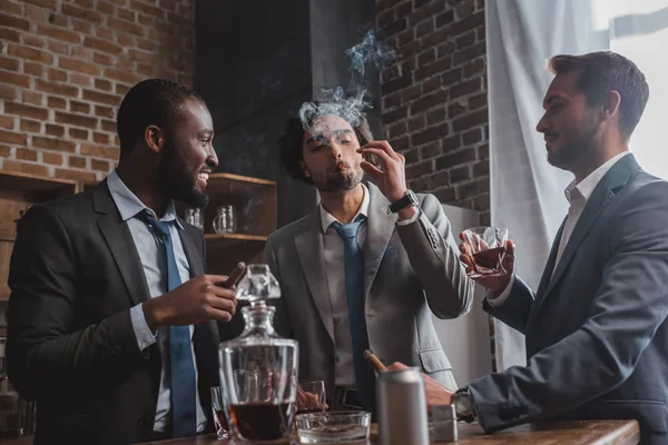 Multiethnic male friends in suits smoking cigars, drinking whiskey and talking — Stock Photo