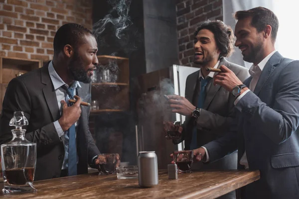 Souriant amis multiethniques masculins en costumes fumer des cigares, boire du whisky et parler — Photo de stock