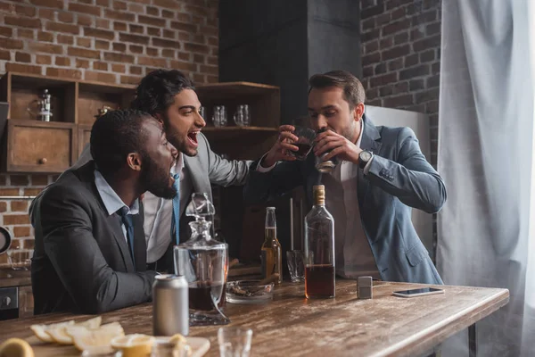 Fröhliche multiethnische Männer, die auf einen Freund blicken, der alkoholische Getränke aus Glas und Dose trinkt — Stockfoto