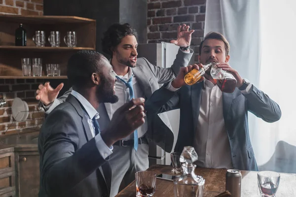 Схвильовані багатоетнічні чоловіки дивляться на друга, який п'є алкогольні напої з пляшок — стокове фото