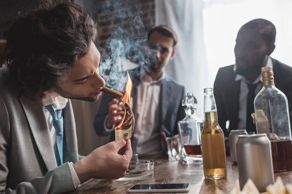 Jovem empresário segurando nota de dólar e fumando charuto enquanto se diverte com os amigos — Fotografia de Stock