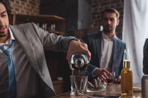 Recortado disparo de hombre en traje vertiendo whisky mientras amigo de pie detrás - foto de stock