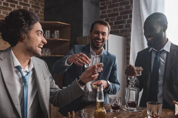 Sonrientes amigos masculinos multiétnicos en trajes bebiendo tequila juntos - foto de stock