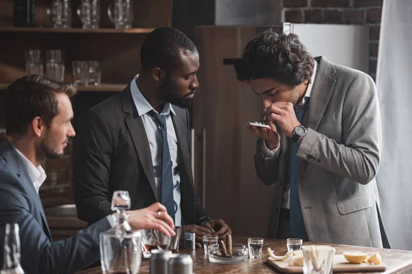 Homens multiétnicos de terno bebendo uísque e olhando para amigo tomando drogas — Fotografia de Stock