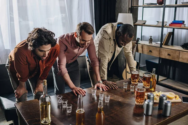 Homens multiétnicos jovens olhando para óculos com bebidas alcoólicas enquanto festejam dentro de casa — Fotografia de Stock