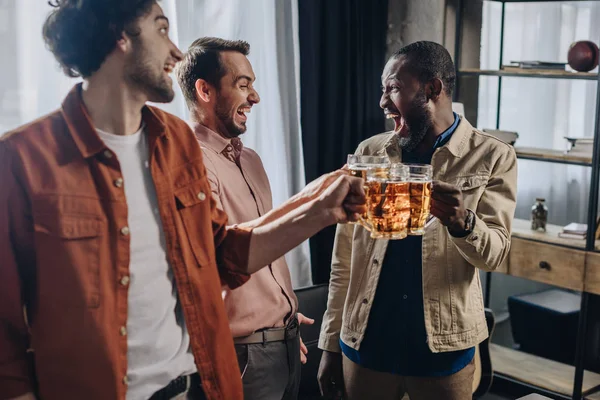 Alegre multiétnico amigos hombres tintineo vasos de cerveza mientras fiesta juntos — Stock Photo