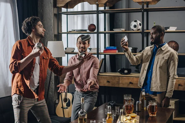 Hombre afroamericano usando smartphone y mirando a amigos haciendo malabares con latas de cerveza - foto de stock