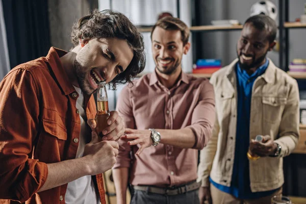 Smiling multiethnic men looking at friend opening beer bottle with teeth — Stock Photo