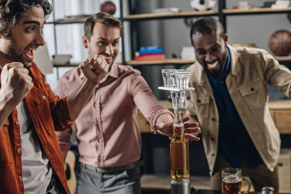 Hombres multiétnicos emocionados construyendo torre de botellas y vasos mientras se divierten juntos en la fiesta - foto de stock