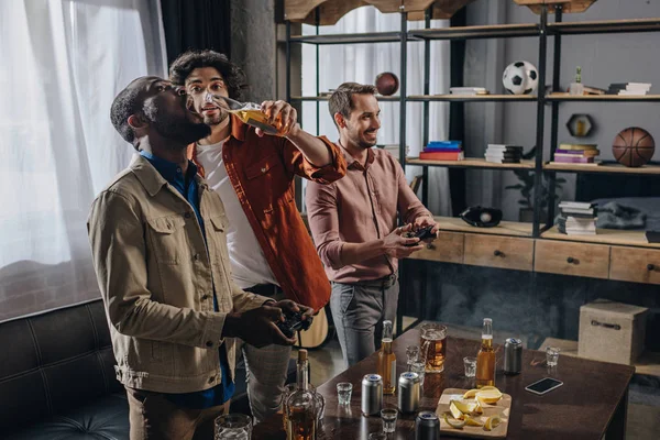 Seitenansicht multiethnischer männlicher Freunde, die Bier trinken und gleichzeitig mit Steuerknüppeln spielen — Stockfoto