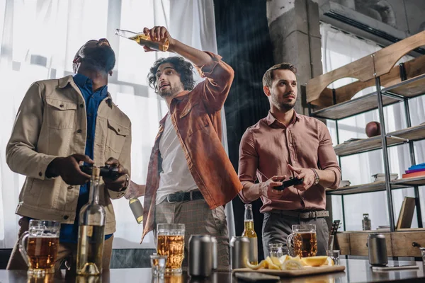 Vista de ángulo bajo de amigos masculinos multiétnicos que beben cerveza mientras juegan con joysticks juntos - foto de stock