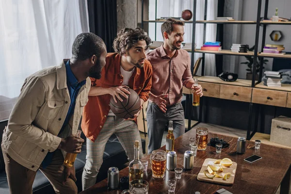 Vista lateral de hombres multiétnicos jugando con pelota de baloncesto y bebiendo cerveza juntos - foto de stock