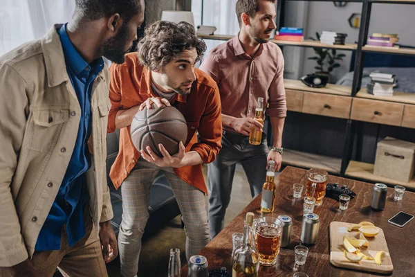 Jóvenes hombres multiétnicos jugando con pelota de baloncesto y bebiendo cerveza juntos - foto de stock