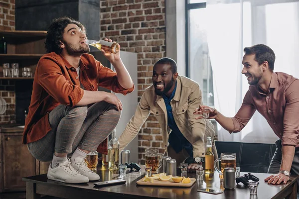 Smiling multiethnic men looking at friend drinking beer while crouching on table — Stock Photo