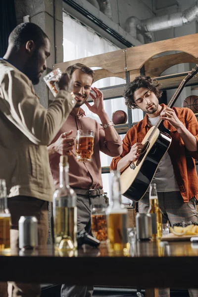 Felices jóvenes amigos multiétnicos beber cerveza y tocar la guitarra juntos - foto de stock