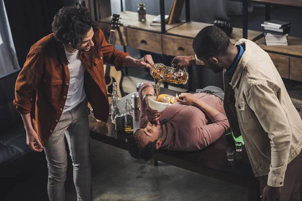 Multiethnic friends pouring beer in funnel and man drinking while lying on table — Stock Photo