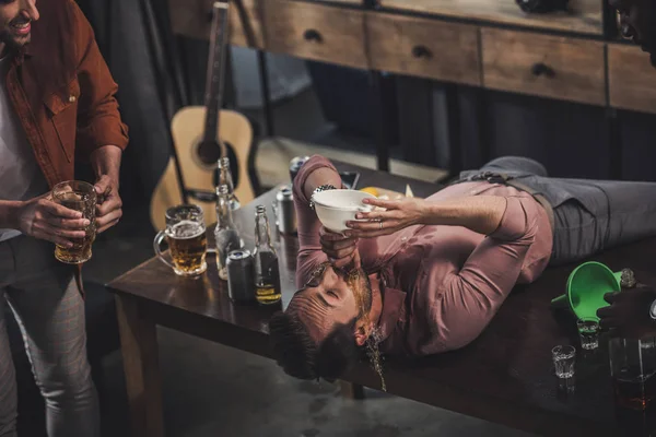 Joven acostado en la mesa y bebiendo cerveza del embudo mientras sus amigos lo miran - foto de stock