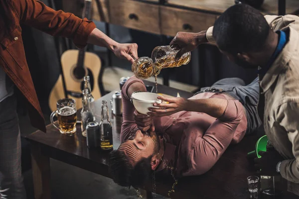 Tiro cortado de amigos derramando cerveja em funil e homem bebendo enquanto deitado na mesa — Stock Photo