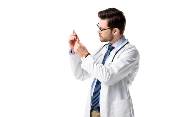 Médico vistiendo bata blanca con estetoscopio y examinando la jeringa aislada en blanco - foto de stock