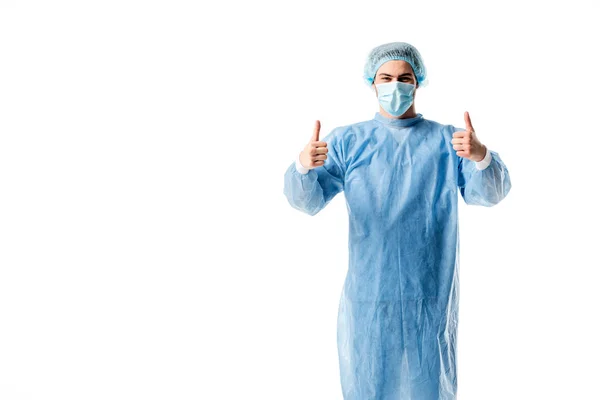 Cirujano vistiendo uniforme azul y gorra médica mostrando pulgares arriba aislados en blanco - foto de stock