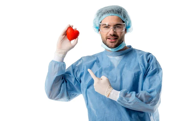 Smiling surgeon in blue medical uniform and medical mask and pointing at toy heart isolated on white — Stock Photo