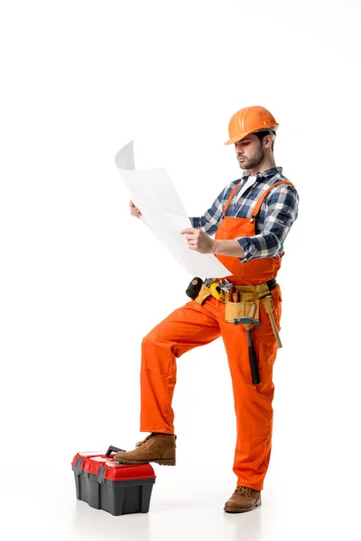 Construtor em laranja geral e capacete olhando para o modelo enquanto se inclina na caixa de ferramentas isolado no branco — Fotografia de Stock