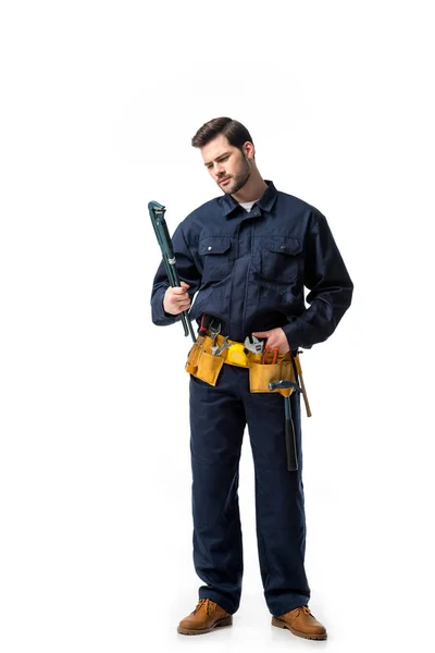 Junger Handwerker in Uniform mit Werkzeuggürtel und Blick auf Schraubenschlüssel isoliert auf weiß — Stockfoto