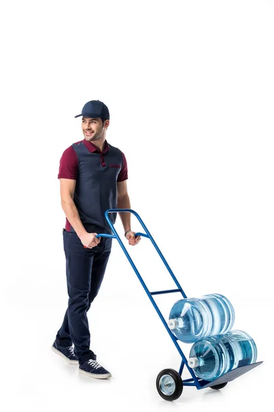 Homem de entrega sorridente em uniforme empurrando caminhão de mão com grandes garrafas de água isolada em branco — Fotografia de Stock