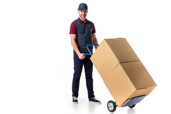 Smiling delivery man in uniform pushing hand truck with cardboard boxes isolated on white — Stock Photo