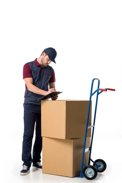 Delivery man in uniform checking notes in notepad with cardboard boxes on hand truck near by isolated on white — Stock Photo