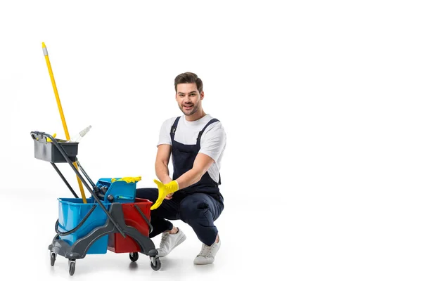 Smiling cleaner in uniform and rubber gloves with cart isolated on white — Stock Photo