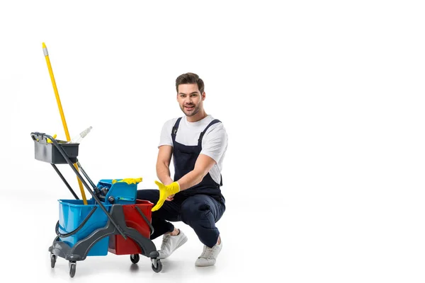 Limpador sorridente em uniforme e luvas de borracha com carrinho isolado em branco — Fotografia de Stock