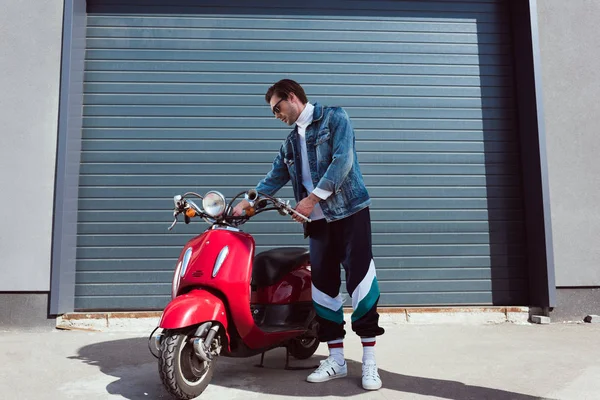 Handsome young man in denim jacket and track pants with vintage red scooter — Stock Photo