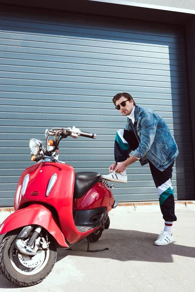 Jovem elegante em roupas vintage com scooter vermelho deitado até atacadores — Fotografia de Stock