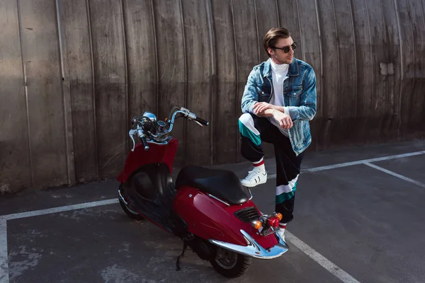 Joven guapo en chaqueta de mezclilla con estilo con scooter vintage en el aparcamiento - foto de stock