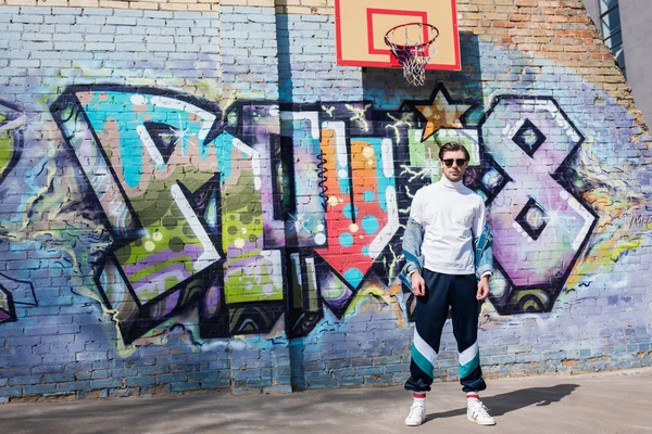 Joven con estilo en ropa vintage delante de la pared de ladrillo con graffiti y anillo de baloncesto - foto de stock