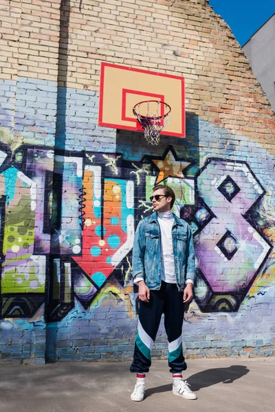 Joven guapo en ropa vintage delante de la pared de ladrillo con graffiti y anillo de baloncesto - foto de stock