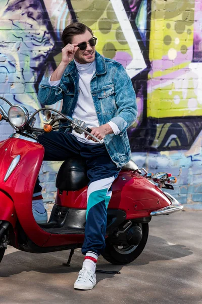 Joven feliz en scooter rojo vintage delante de la pared de ladrillo con graffiti - foto de stock