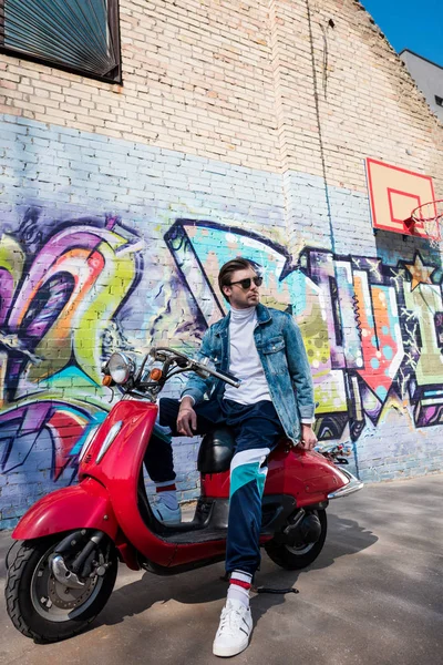 Красивий молодий чоловік у стильному одязі на старовинному червоному скутері перед цегляною стіною з графіті — стокове фото