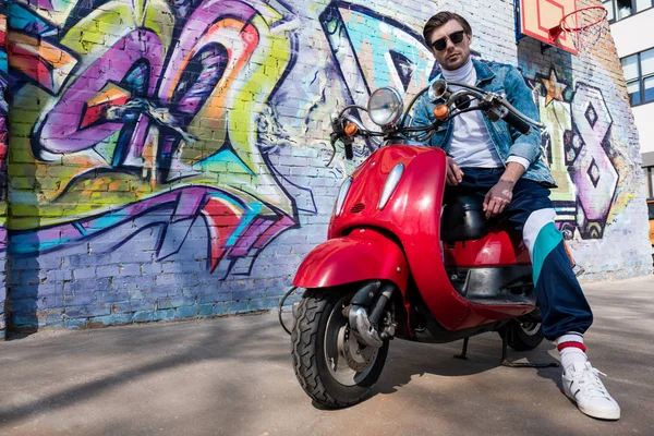 Jovem bonito na scooter vermelho vintage olhando para a câmera na frente da parede de tijolo com graffiti — Fotografia de Stock