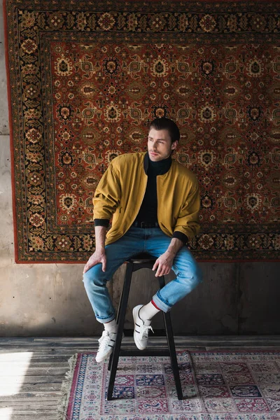 Stylish young man sitting on chair in front of rug hanging on wall — Stock Photo